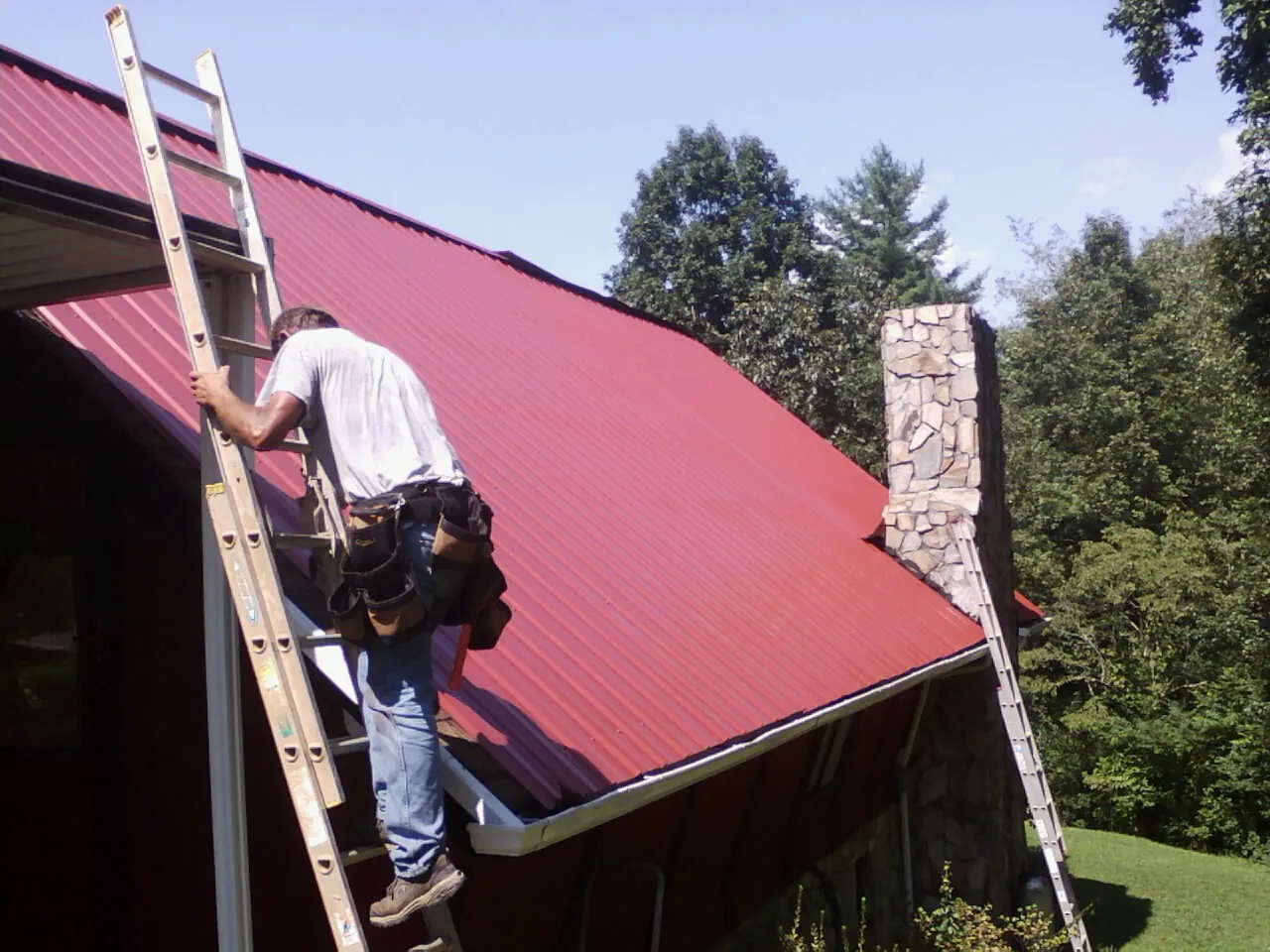 Roofer on a ladder beside a red metal roof and stone chimney — Soffit & Fascia Repair by Roofing Brothers in Jarrell